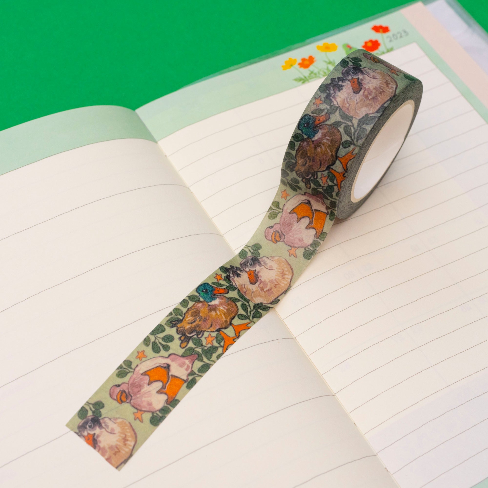the image shows a roll of washi tape with a charming design featuring ducks and floral elements, unfurling across the pages of an open notebook. the notebook has lined pages and a light green cover with subtle floral accents. the background is a vibrant solid green. the overall aesthetic is soft, pastel, and charming, suggesting a crafting or journaling setting. the tape itself appears to be highquality and aesthetically pleasing, with soft colors and delicate designs.