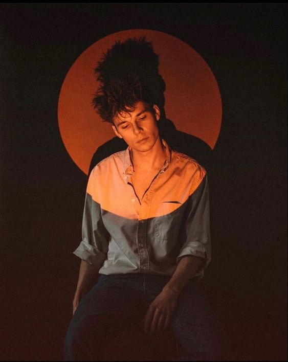 a young man with dark, tousled hair is captured in a moody portrait. he sits against a black backdrop, illuminated by a circular orange spotlight that creates a halo effect around his head and torso. he wears a light, collared shirt, halftucked into dark jeans, and gazes downward with a contemplative expression. the overall atmosphere is artistic and introspective, hinting at themes of music, creativity, or emotional depth.