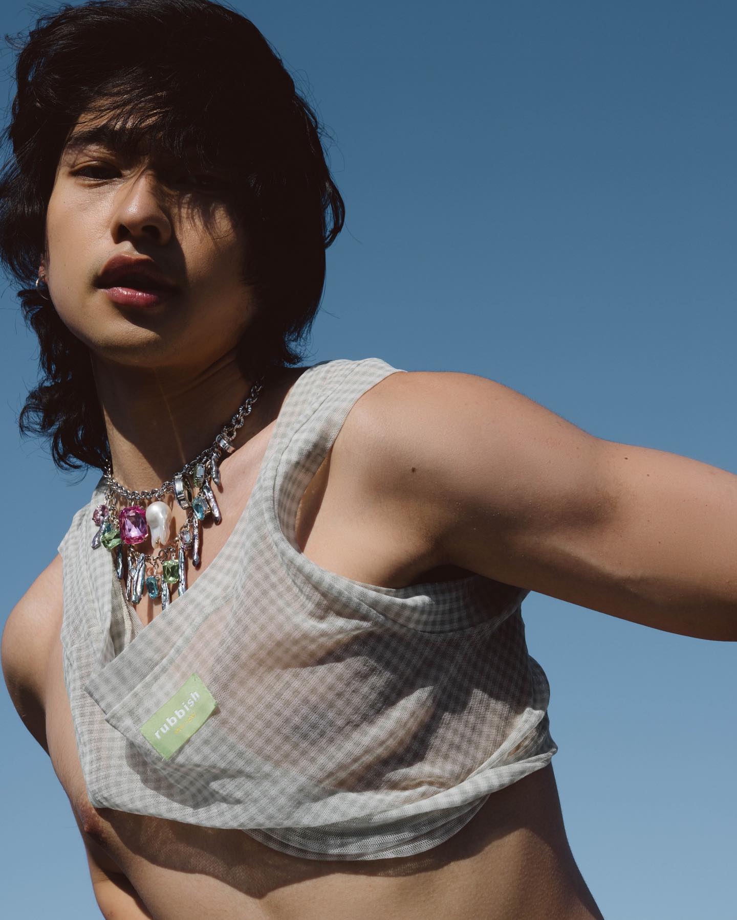 a young adult male with mediumlength black hair styles a trendy androgynous look. he wears a sheer, gingham cropped tank top and a statement silver necklace featuring an eclectic mix of colorful charms and a large pearl.  the image is a lowangle shot against a bright, clear blue sky, emphasizing the model's confident pose. the sunlight illuminates the scene, highlighting the textures and details of the clothing and jewelry.