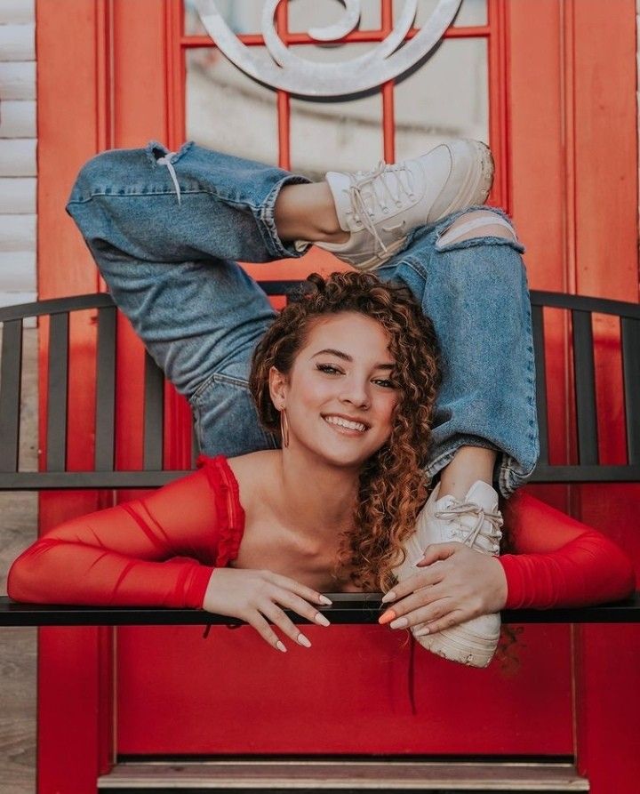 a young woman with curly hair is sitting on a bench in front of a red door, she is bending her legs and arms into a flexible pose, smiling at the camera. she is wearing a red top and ripped jeans and white shoes. her nails are painted in a bright color.