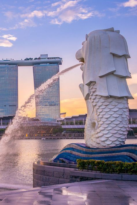 the image showcases the iconic merlion statue in singapore at sunset.  a large fountain sprays water from the merlion's mouth, creating a dramatic effect against the backdrop of the marina bay sands hotel. the sky displays soft pastel hues of pink and orange, complementing the white of the statue and the modern glass architecture of the hotel. the scene is tranquil yet dynamic, highlighting both the natural beauty and manmade marvels of singapore. the foreground shows the paved area around the statue, adding depth to the overall composition.
