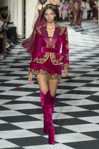 a model walks the runway in a stunning fuchsia velvet ensemble. the outfit consists of a cropped, embroidered velvet blazer and matching shorts, adorned with intricate gold detailing. the model is also wearing a matching pair of high, fuchsia velvet boots. the overall style is elegant, glamorous, and luxurious, reflective of highfashion runway attire.  the background features a black and white checkered floor and other models can be seen in the distance, blurred.