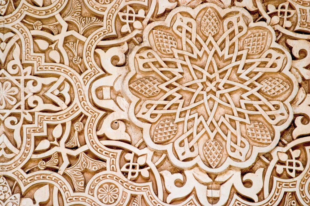 the image shows a detailed closeup of an intricately carved wall decoration, likely plasterwork or stucco, featuring complex islamic art patterns. the patterns consist of geometric and floral motifs, including a prominent rosette design with interlacing ribbons and textured petallike shapes. the play of light and shadow emphasizes the relief carving, creating a textured and ornate surface. the style is characteristic of moorish architecture, possibly found in the alhambra palace.