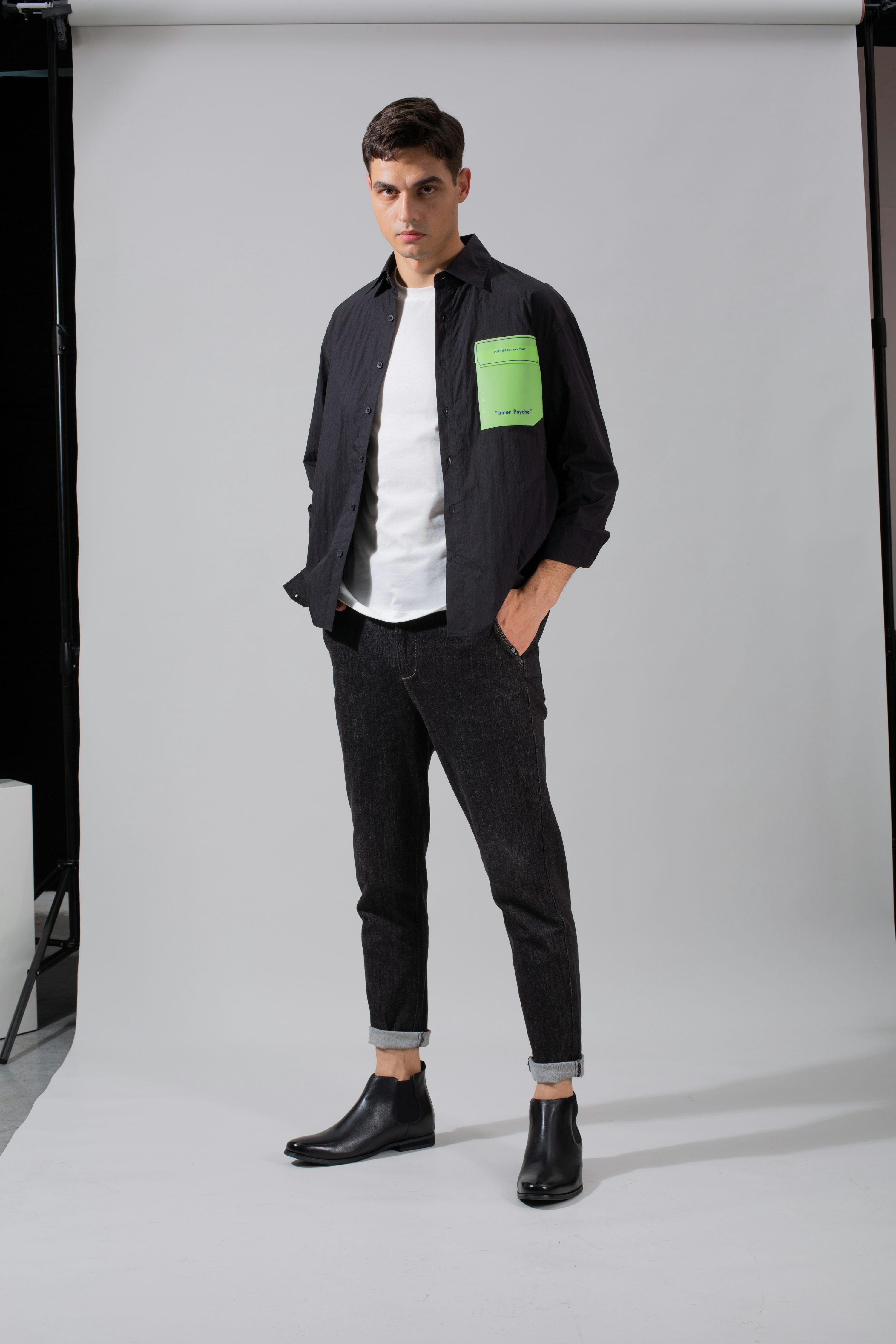 a fullbody studio shot of a male fashion model. he is wearing a black buttondown shirt with a lime green rectangular patch on the left chest, dark wash jeans, and black chelsea boots. the model is standing against a plain white background, and his pose is relaxed yet stylish.  the overall aesthetic is clean, minimalist and contemporary.