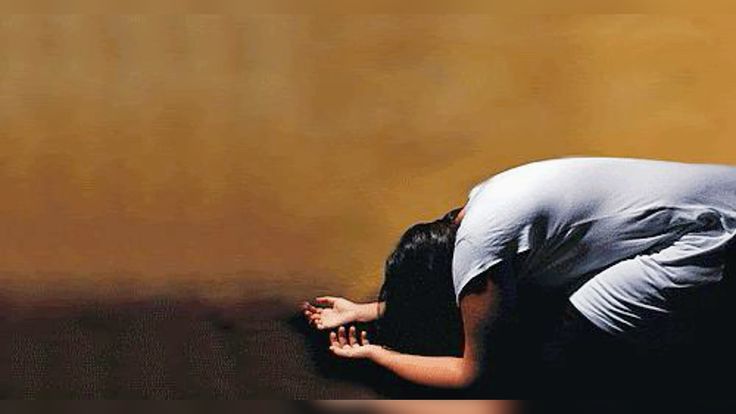 a woman is kneeling on the floor with her hands together in prayer. the background is a simple golden color, providing a serene setting. the woman's posture and expression convey a sense of deep devotion and prayer.