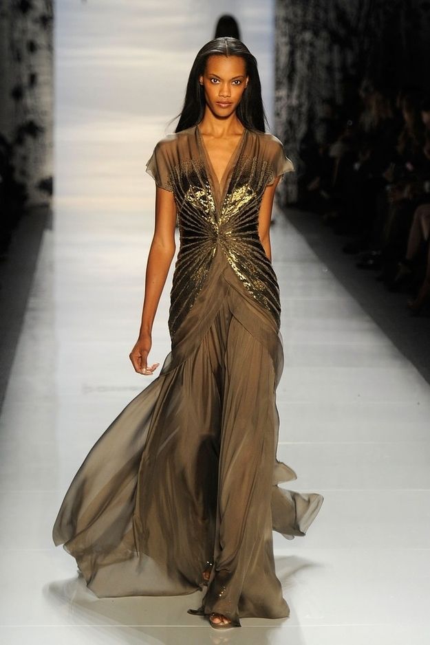 a black woman model with long dark hair walks down a runway in a long, flowing brown dress with gold embellishments. she is posing for the camera with a sophisticated and elegant demeanor. the dress is very flattering on her body, and she looks absolutely stunning.