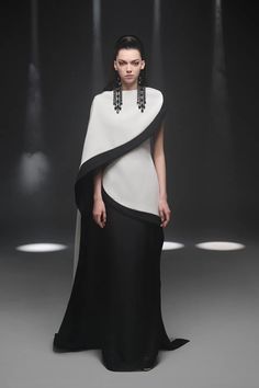a fashion model walks down a runway wearing a white and black cape dress with a long skirt and a black top. she is posing confidently for the camera, and her dress is flowing gracefully behind her. the studio lighting is highlighting her elegant and sophisticated look.