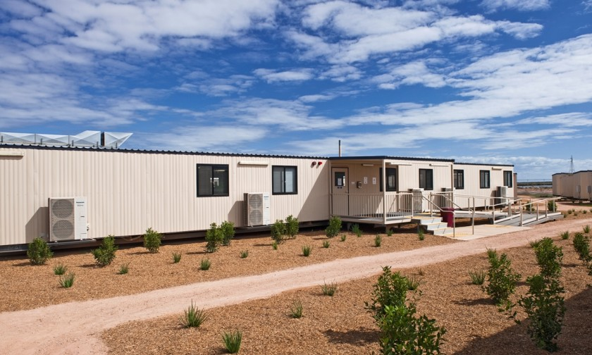 a beige colored modular building with a black roof and windows, with a covered walkway. the building sits on a gravel path, with dry, brown grasses and shrubs in the foreground. the sky is blue with fluffy, white clouds.