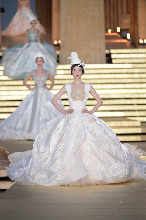 a model walks the runway in a stunning white wedding gown. the dress is intricate and luxurious, featuring delicate lace, embroidery, and a flowing silhouette. the model is styled with a white hat and minimal makeup, showcasing the beauty of the dress. the background is a blurred image of a stage and audience, creating a sense of excitement and anticipation. the overall image is a celebration of fashion, beauty, and elegance.