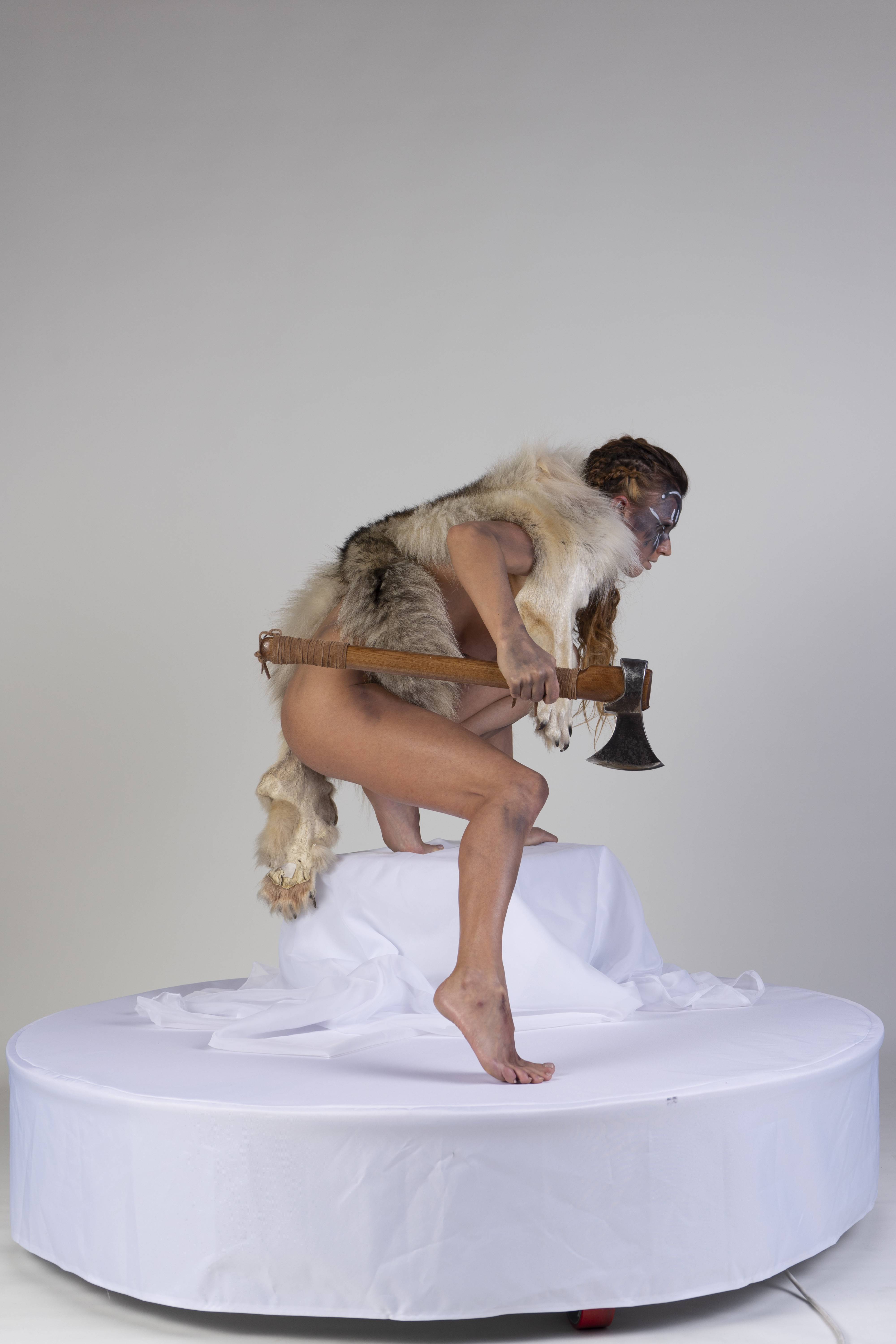 a nude woman stands on a white platform, wearing a wolf pelt draped over her shoulders and carrying an axe. she has goggles on and face paint around her eyes. she has braids in her hair. the background is a plain white studio.
