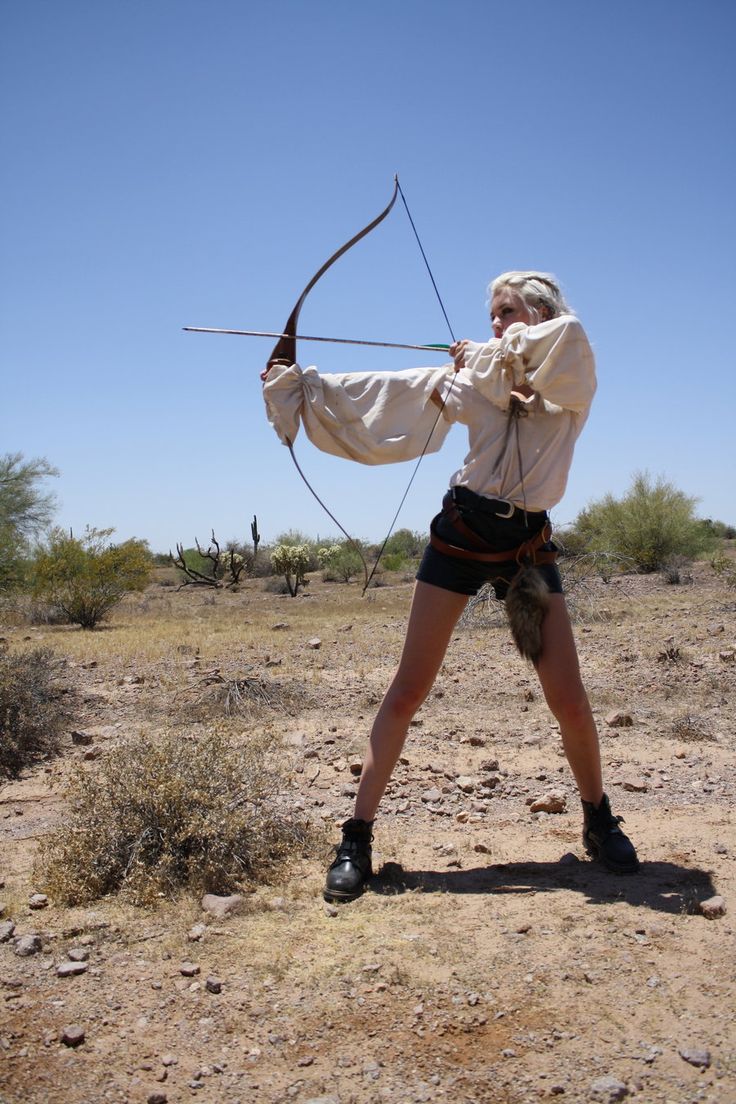 a woman in a medieval inspired costume is holding a bow and arrow in the desert. she is aiming at a target offcamera. she is wearing a white tunic with a dark belt and dark shorts.