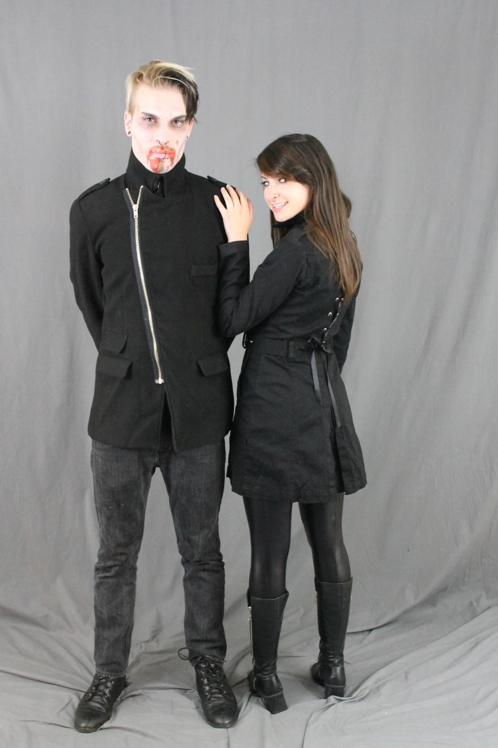 full shot of a man and woman dressed in dark, gothicstyle clothing. the man is styled as a vampire, complete with pale makeup and fake blood. the woman is wearing a black coat. both are standing against a gray background.