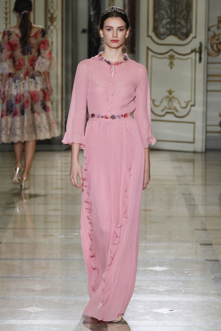 a full shot of a model gracefully walking down a runway, showcasing a long, pleated, light pink dress with delicate floral details around the waist. the dress has threequarter sleeves and a subtle ruffle detailing along the skirt. the model is elegantly posed, her hair pulled back, and her makeup is natural. the background features a marble floor and an ornate interior with large doors, setting a highfashion atmosphere. another model in a floral dress is visible in the background, out of focus. the overall image projects a sense of sophistication and elegance.