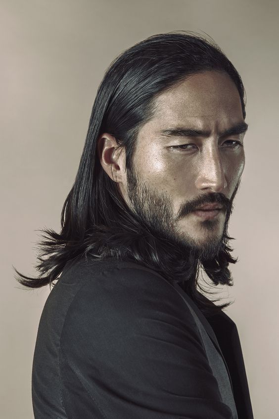 a portrait of a seriouslooking asian man with long, dark hair and facial hair (beard and mustache) wearing a black jacket in a studio setting.