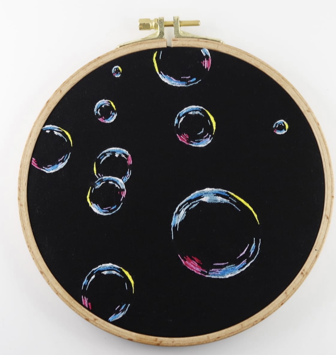 a closeup shot of a black embroidery hoop with colorful bubble designs on a black background. the embroidery uses various colored threads, creating a colorful and playful design. the edges of the hoop are visible, giving the image a crafty feel.