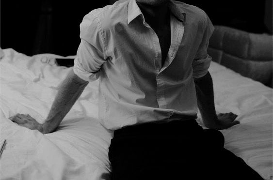 a man in a white shirt sits on the edge of a bed, leaning on his forearms. he has his legs tucked to the side of his torso, as if he's sitting on the edge of a bed. the man's chest is partially exposed with a slight view of his upper torso, and his legs are out of view. his shirt is white, unbuttoned at the top, and his pants are black. the image is in grayscale, with only the man and the bed visible, with dark shadows behind him.