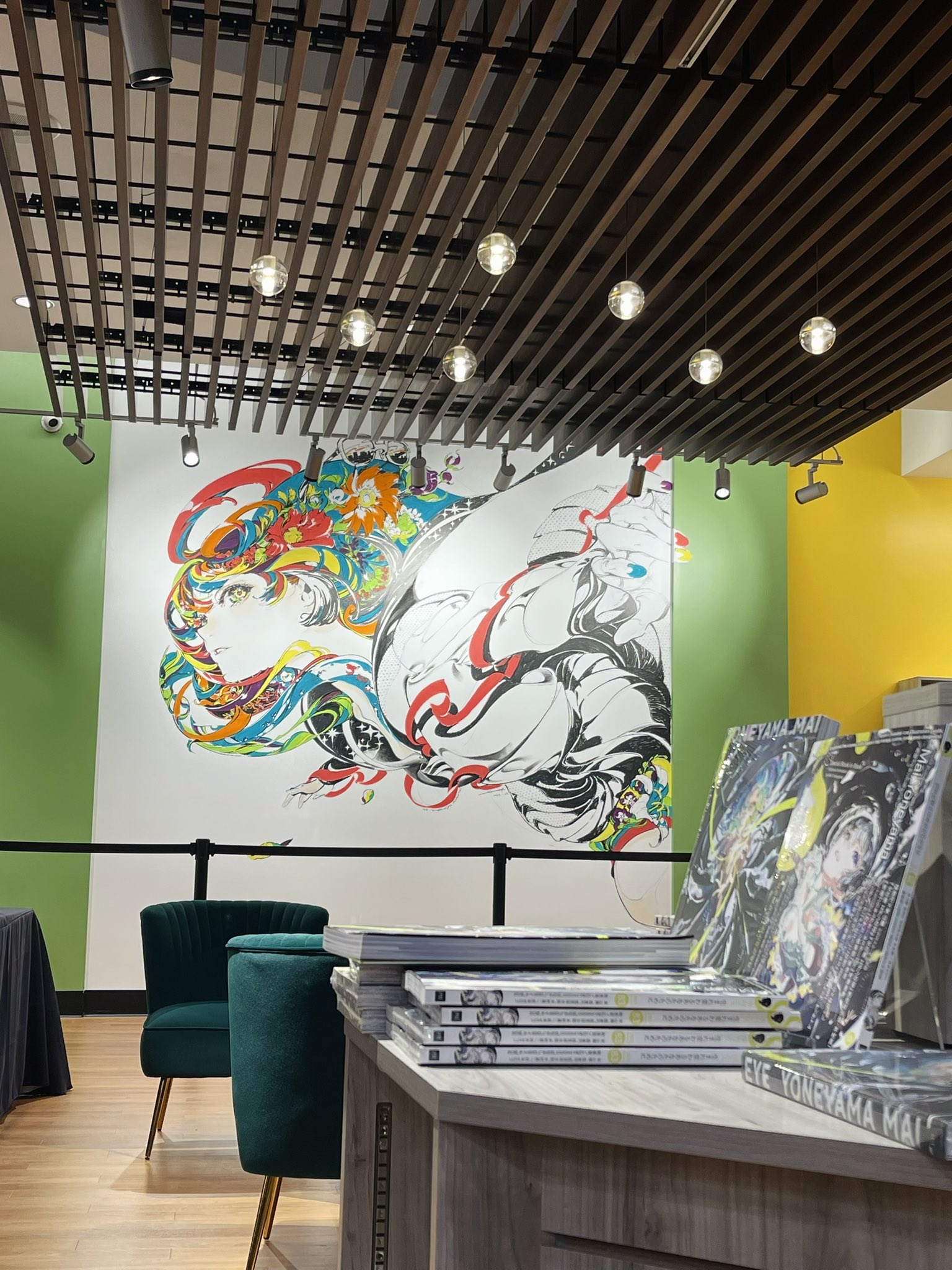 the image shows the interior of a bookstore with a large art piece on the left wall. the art piece depicts a woman's face in profile, surrounded by colorful and abstract designs. the artwork is in black and white with pops of red, blue, and yellow. in front of the artwork are two green velvet chairs. to the right of the artwork is a display of art books and manga. the ceiling of the store is black with wooden beams and recessed lighting. the walls are painted green and yellow.