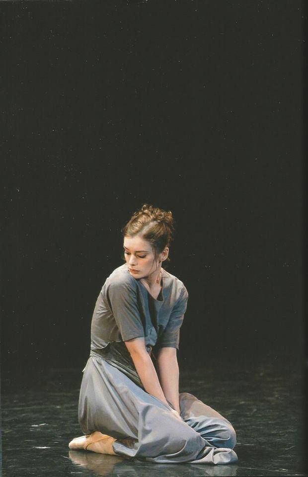 a lone female ballerina dressed in a simple gray dress sits gracefully on a dark stage. her posture conveys a sense of focused emotion, with her arms crossed and her gaze directed downwards. the dramatic lighting casts a spotlight on her, emphasizing her isolation and the intensity of her performance. the minimalist backdrop of the stage allows her artistry to shine through, highlighting her elegance and the graceful lines of her body.