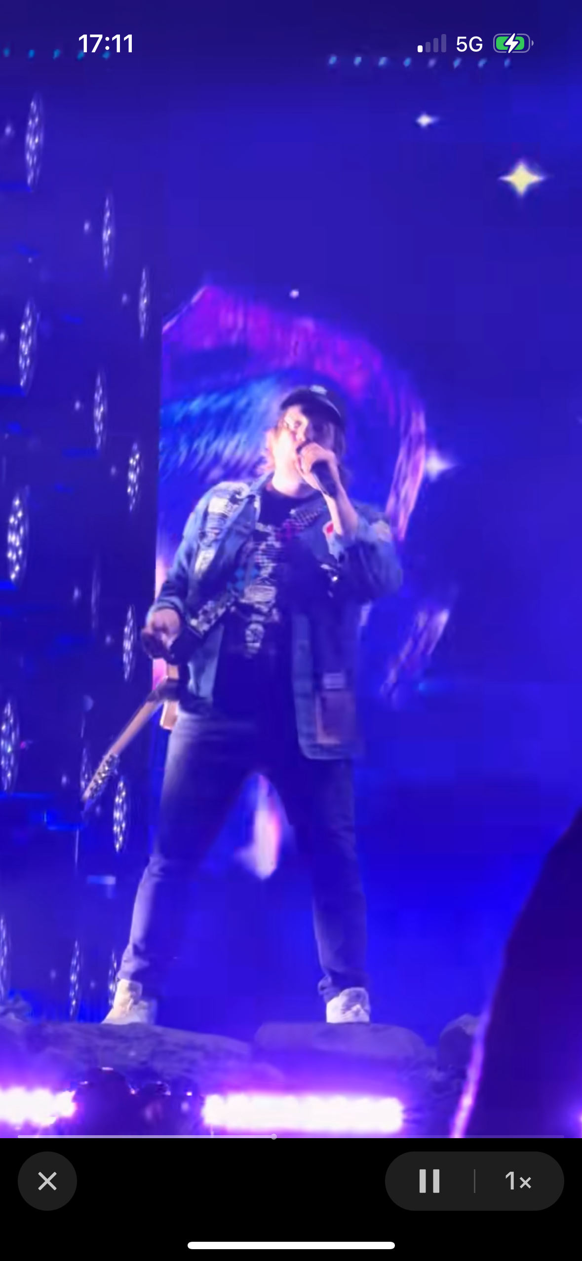 a singer performs on a stage with purple and blue lights. they hold a microphone with one hand and what looks to be a guitar in the other. the stage looks to be lit up in the background. the stage looks to be lit up in the background. there is a blur of people to the right of the musician.