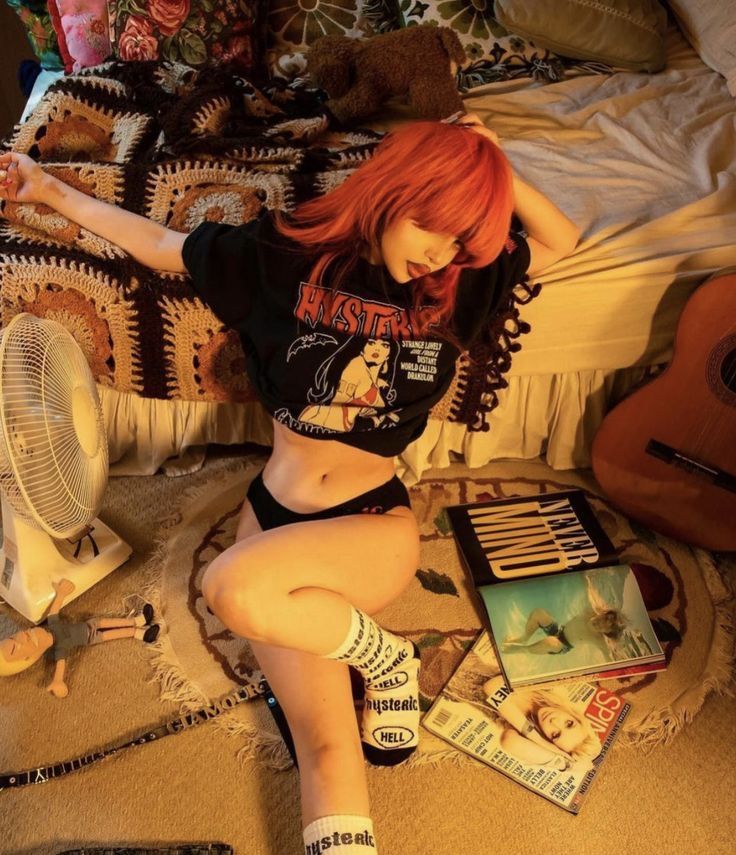 the image shows a young woman with bright orange, short, straight hair and bangs. she's wearing a black hysteric glamour tshirt featuring a vintage graphic, black underwear, and white socks with black lettering. she's sitting on a bed with a vintage crocheted blanket, a classic acoustic guitar, a box fan, and several magazines including nirvana's nevermind and a spice girls magazine, scattered around her. the overall aesthetic is retro, suggesting a 70s or 80s vibe, with a focus on vintage clothing and rock and roll culture. the lighting is warm and adds to the nostalgic atmosphere.