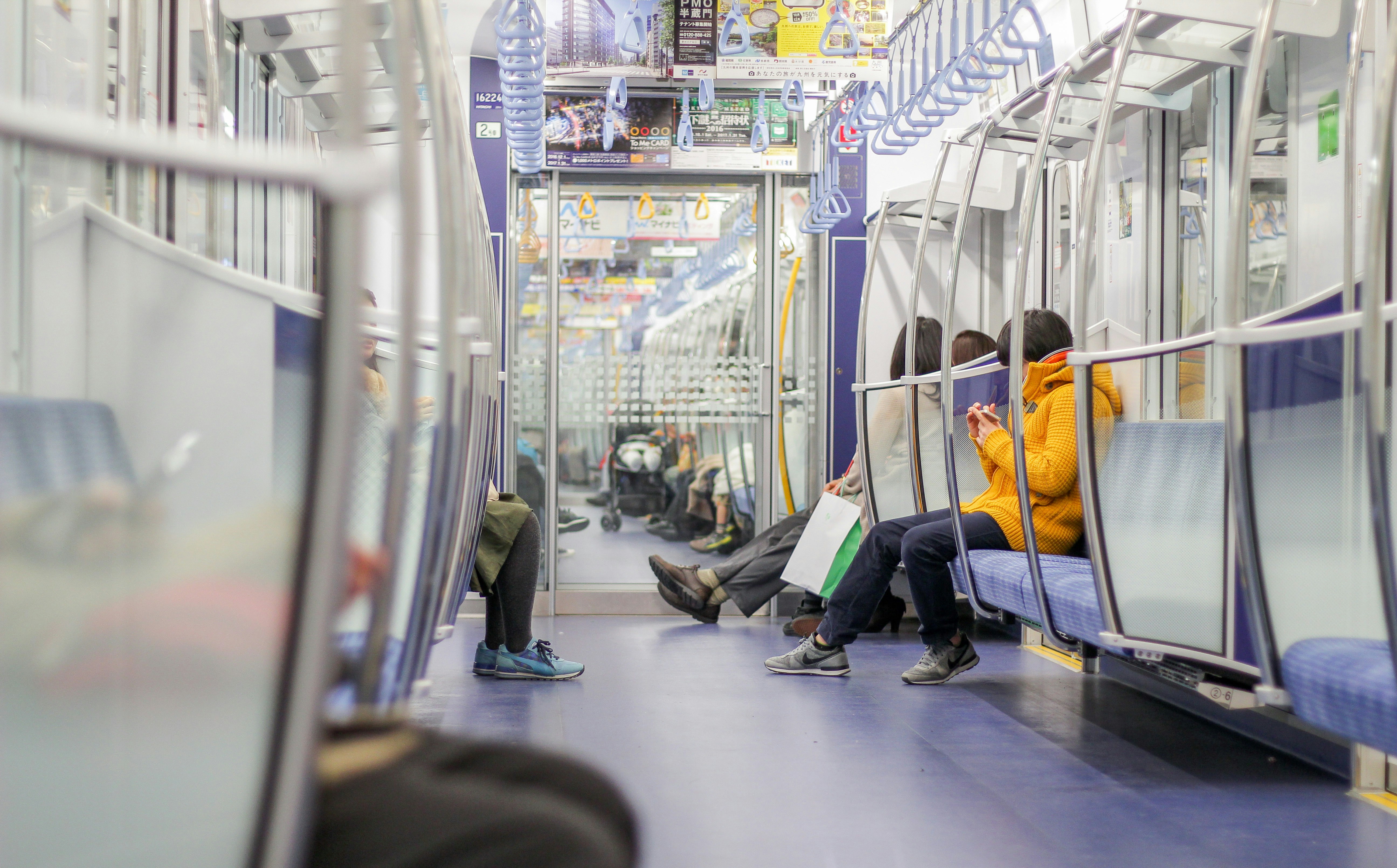 an interior shot of a train carriage showing passengers seated on blue upholstered benches. the train car features silver handrails, overhead advertisements, and reflective windows. some passengers are visible, including one wearing a bright yellow sweater. the floor is a dark blue color.