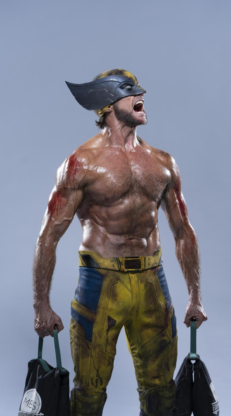 a man dressed as wolverine, with a muscular physique, scars, and wearing the character's signature mask and yellow and blue costume. he is yelling and holding two black bags.