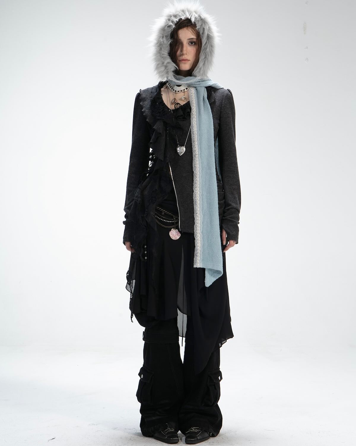 fullbody shot of a woman model showcasing a stylish outfit. she's wearing a gray furlined hood, a light blue scarf, a black jacket with ruffles, a black sheer overlay skirt, and black pants with intricate details around the ankles. she accessorizes with a necklace and has dark boots. the backdrop is a simple white studio setting. the overall style evokes a blend of goth and bohemian aesthetics.