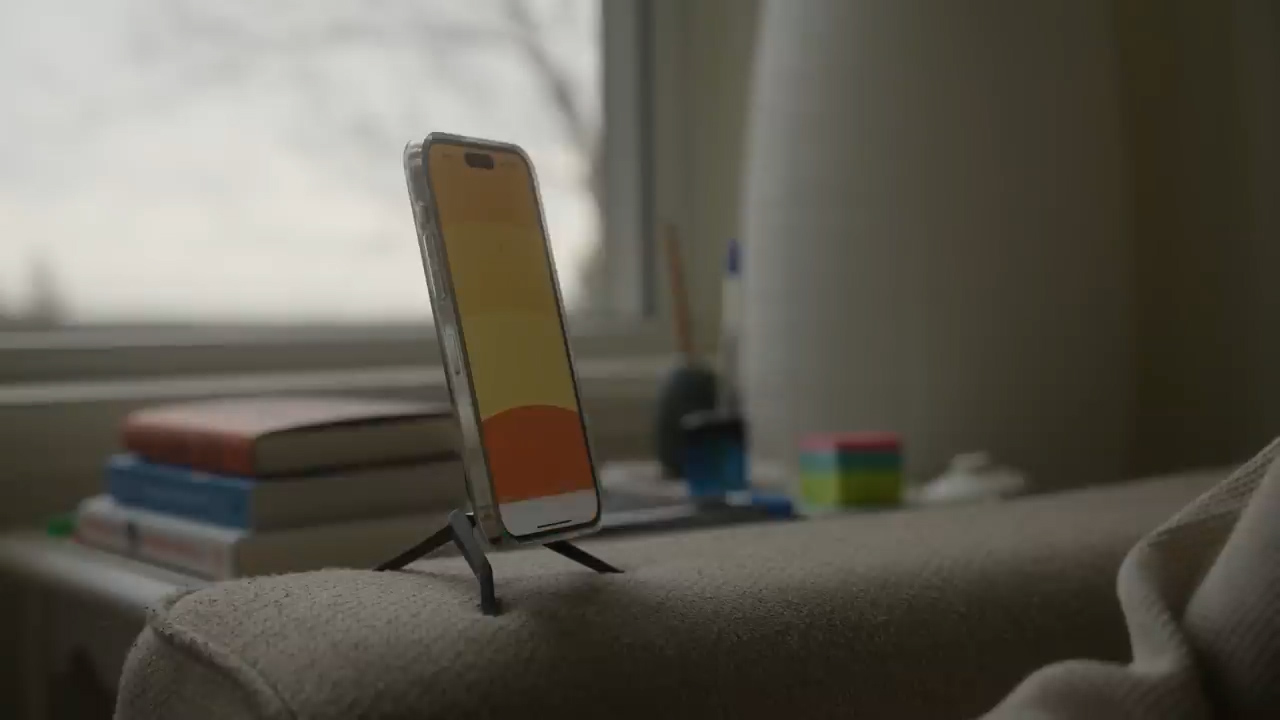 a smartphone rests on a small stand on the arm of a couch. the phone displays a warmtoned image, possibly a sunset. in the background, there's a table with a stack of books and writing utensils. the scene is bathed in soft, natural light, suggesting a relaxing atmosphere.