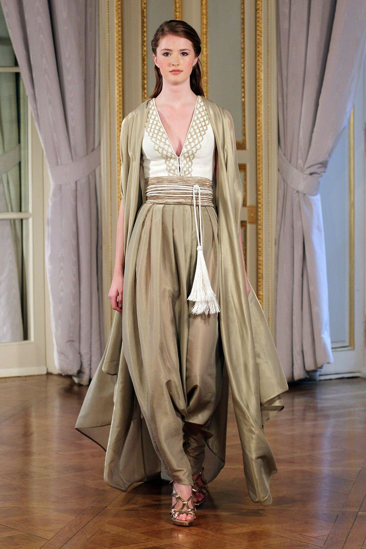 a woman is walking on a runway in a fashion show. she is wearing a white top with a gold pattern around the neckline, tan trousers with a large tassel detail, sandals, and a tan cape. the background is a room with curtains and gold trim.