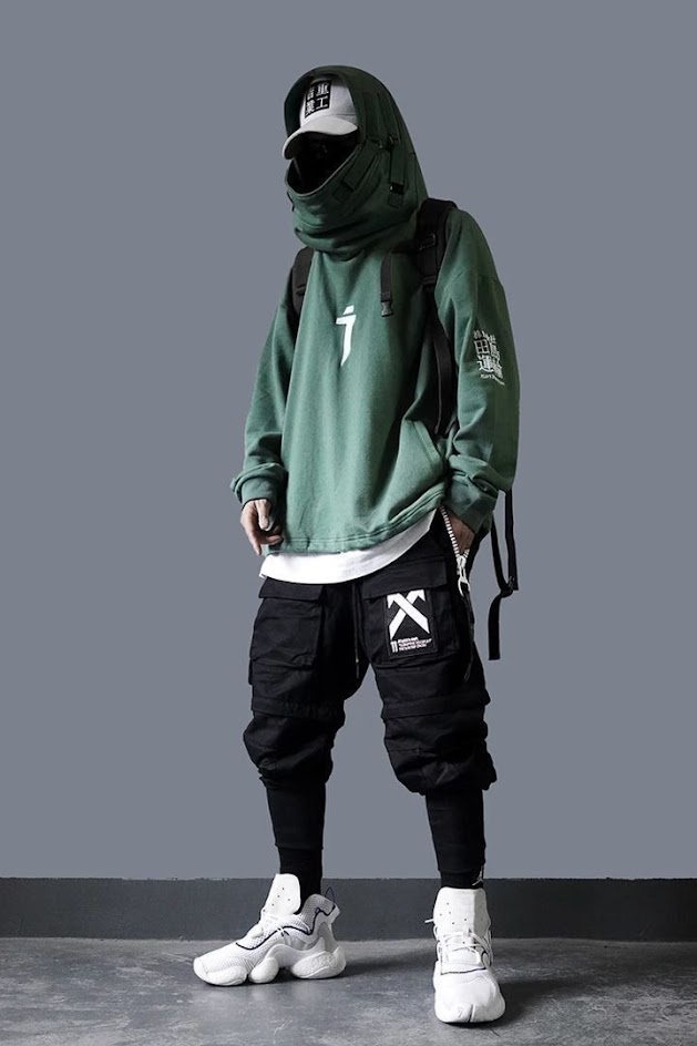 a man is wearing a green hooded sweatshirt, black cargo pants, white sneakers, and a white cap. he is also wearing a black backpack. he is standing in front of a gray background.
