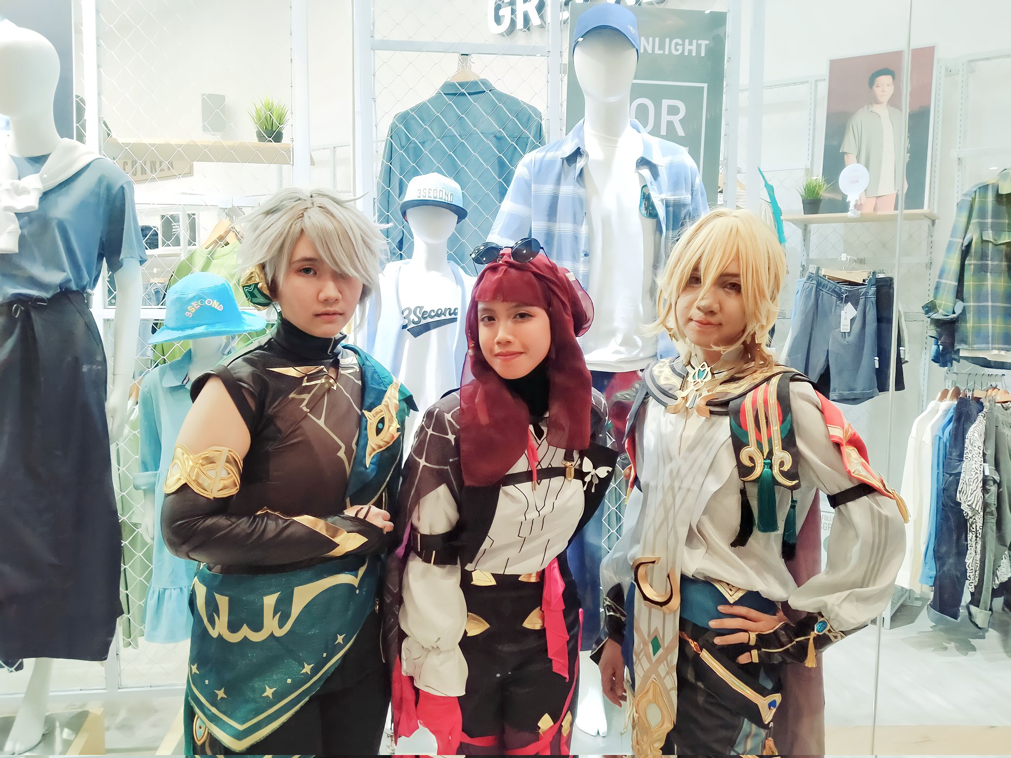 three young people in cosplay, dressed as anime characters, posing in front of a store display. they're dressed in colorful, elaborate costumes with props and accessories. two of them are female, and one is male.