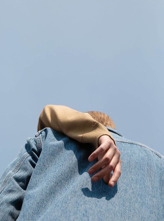 a low angle shot of two people embracing with one person wearing a tan sweater and the other person wearing a blue denim jacket, standing against a bright blue sky. the person in the tan sweater has their hand resting on the shoulder of the person in the denim jacket.  the photo captures a feeling of love, affection, and togetherness.