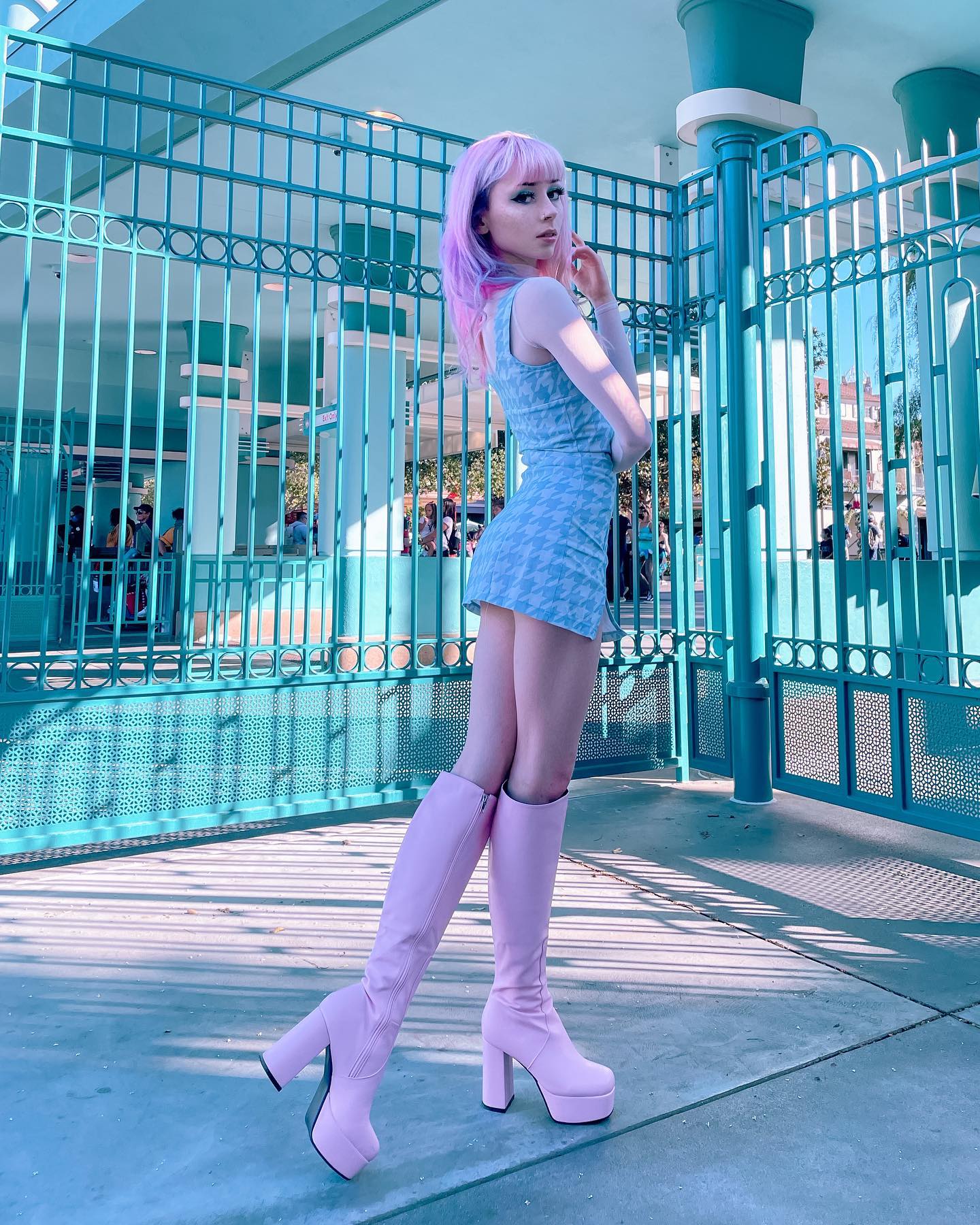 a young woman with pastel pink hair is standing in front of a teal metal fence. she is wearing a light blue mini dress and light pink platform kneehigh boots. the setting appears to be outdoors on a sunny day.  her pose is stylish and confident. the overall aesthetic is retro and pasteltoned.