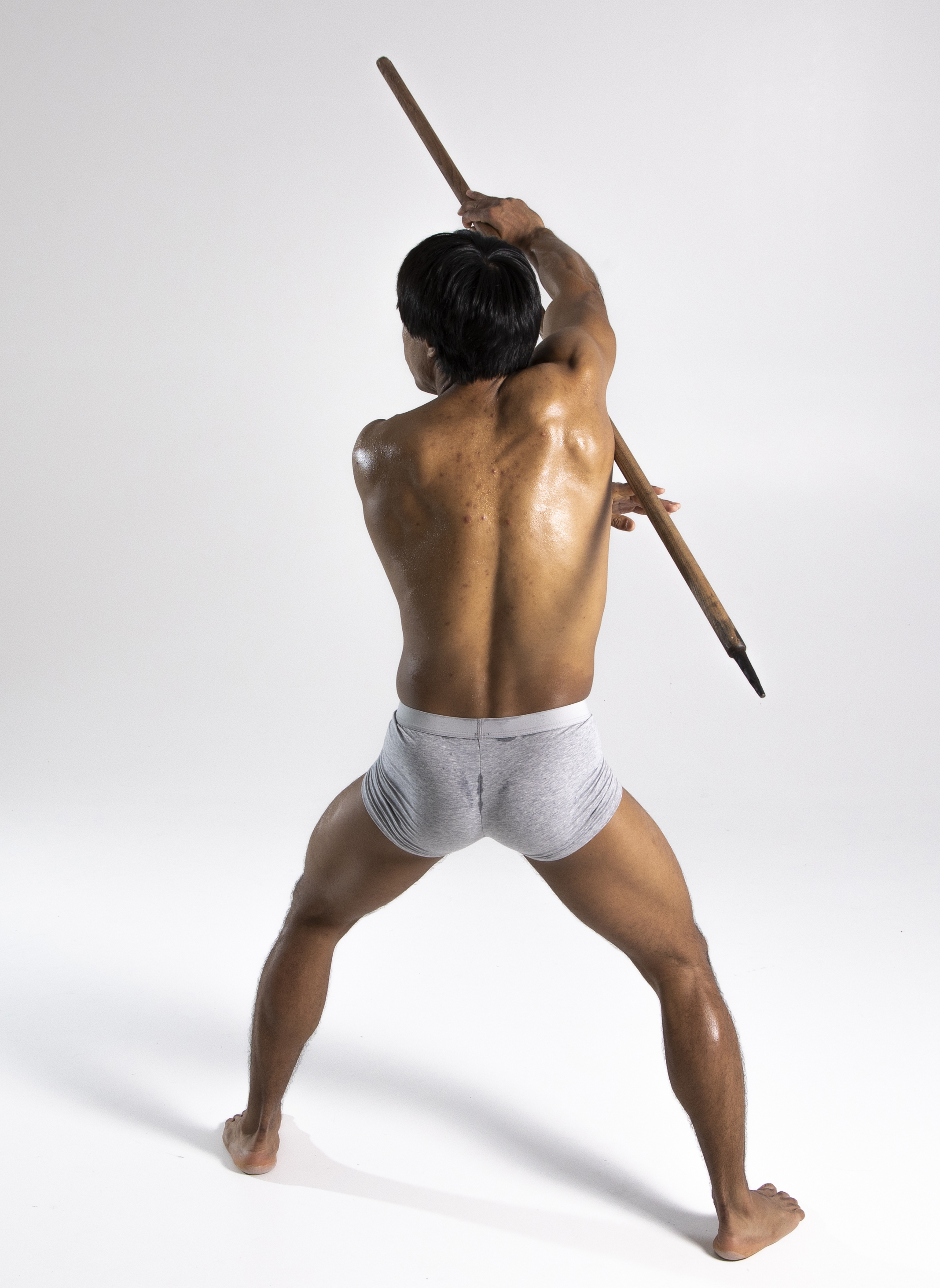 a shirtless man with dark hair is seen from the back, standing with his legs spread apart and holding a long wooden pole over his right shoulder. he is wearing gray underwear and is set against a plain white background in what appears to be a studio setting. his physique suggests he may be an athlete or someone who exercises regularly.