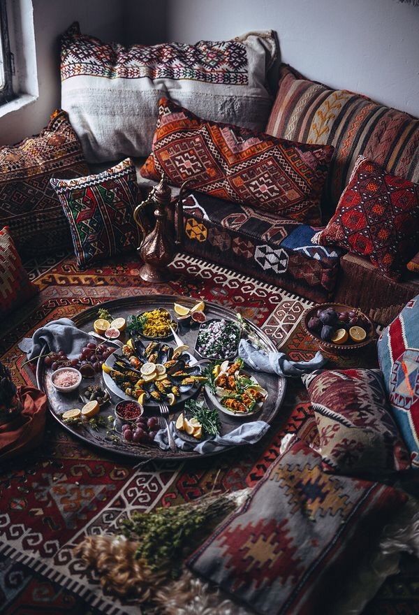 the image showcases a lavish spread of traditional turkish food arranged on a large round silver tray. the tray is filled with various dishes including mussels with lemons, rice, salads, and fruits like grapes and sliced lemons. small bowls of salt and red pepper flakes are also visible. the tray is placed on a richly patterned rug surrounded by decorative cushions and pillows, creating a cozy, orientalstyle setting. a turkish coffee pot sits among the cushions, and a window is visible in the corner. the overall scene suggests a relaxed, informal dining experience.