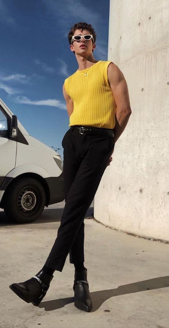 a man wearing sunglasses, a yellow tank top, black pants, and boots stands in front of a white van next to a concrete wall. he is looking away from the camera.