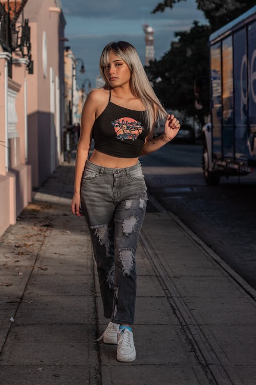 a young woman with blonde hair and bangs is walking down a city street. she is wearing a black crop top with a graphic design, grey ripped jeans, and white sneakers.  the photo is taken from a slightly low angle and shows her from the knees up.  the setting appears to be an urban area with buildings in the background and a truck parked on the side of the street. the lighting suggests it's likely daytime. the overall feel of the photo is stylish and casual.