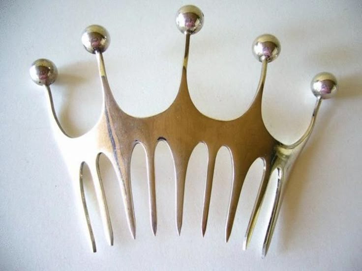 a closeup shot of a decorative silver metal comb shaped like a crown. the comb has five rounded points at the top, each topped with a silver ball. the lower part of the comb has several teeth. the comb is on a lightcolored surface.