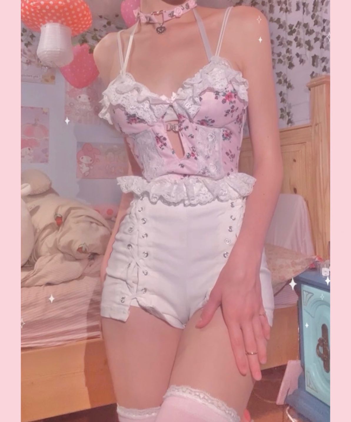 the image shows a young woman wearing a charming and delicate pink floral corset top with white lace trim, paired with highwaisted white shorts that have laceup detailing on the sides. she also wears a heartshaped choker necklace and pink thighhigh stockings with lace at the top. the setting appears to be a bedroom, with soft pink lighting and pastelcolored decor, including mushroomshaped lamps in the background, enhancing the overall sweet and romantic aesthetic. the overall style is very kawaii and girly.