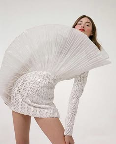 a young woman in a white, pleated, lace mini dress with long sleeves, poses for the camera. the dress is styled with a fanlike shape that appears to be a part of the dress. the model is standing in front of a white background.