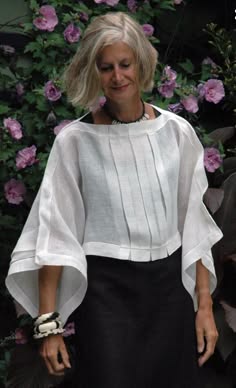 a mature woman with gray hair, is standing outdoors wearing a long white blouse with black skirt and a white bracelet, she is standing in front of a bush full of pink flowers.