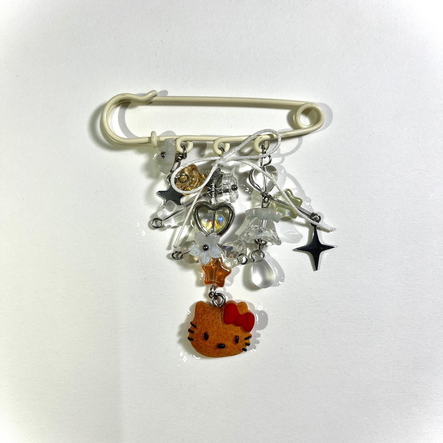 a closeup shot of a unique handmade brooch. the base is an offwhite colored safety pin from which various charms are suspended. these include stars, beads, clear acrylic shapes (like flower petals and water droplets), and a heart. at the bottom, there's a hello kitty charm with a red bow. the overall aesthetic is kawaii and decorative.