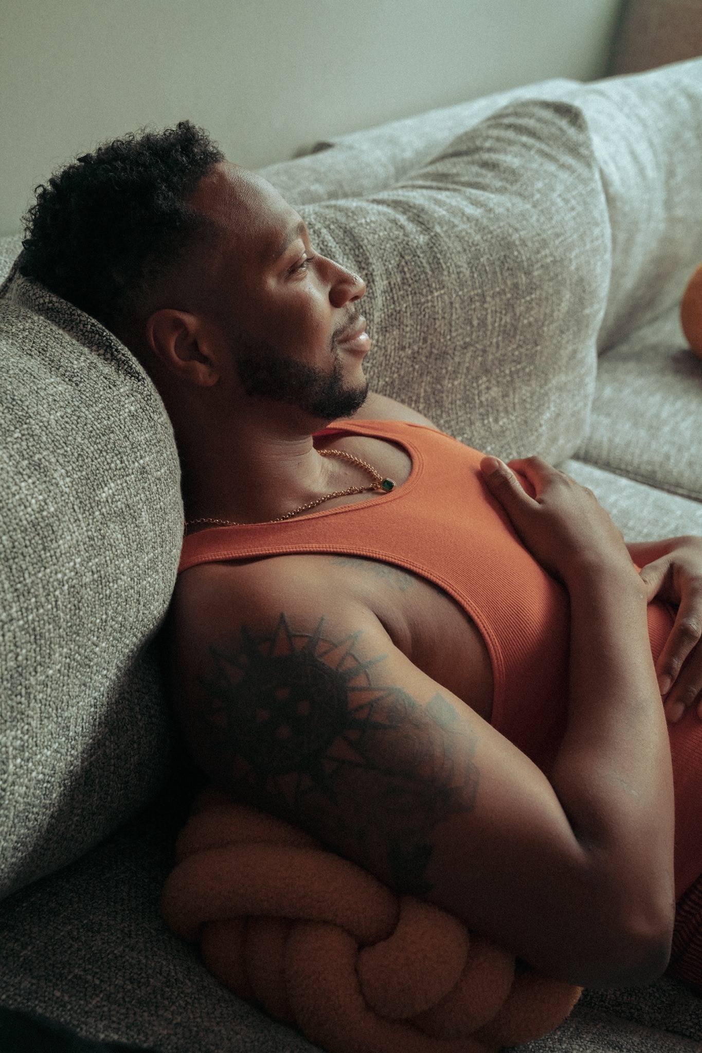 a man in an orange tank top lays on a couch, with a pillow under his head, relaxed with eyes closed. he has a tattoo on his left shoulder. the couch is covered in a gray fabric.