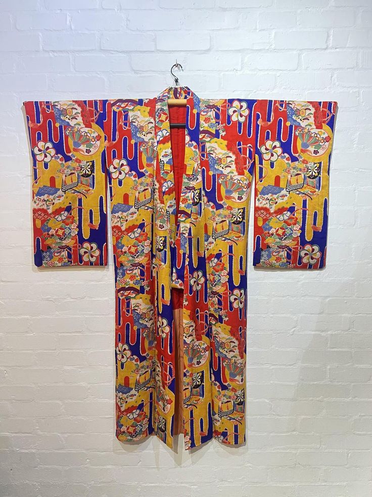 a colorful kimono hangs against a white brick wall. the kimono is red, yellow, and blue with a floral pattern. it is hanging on a hanger.