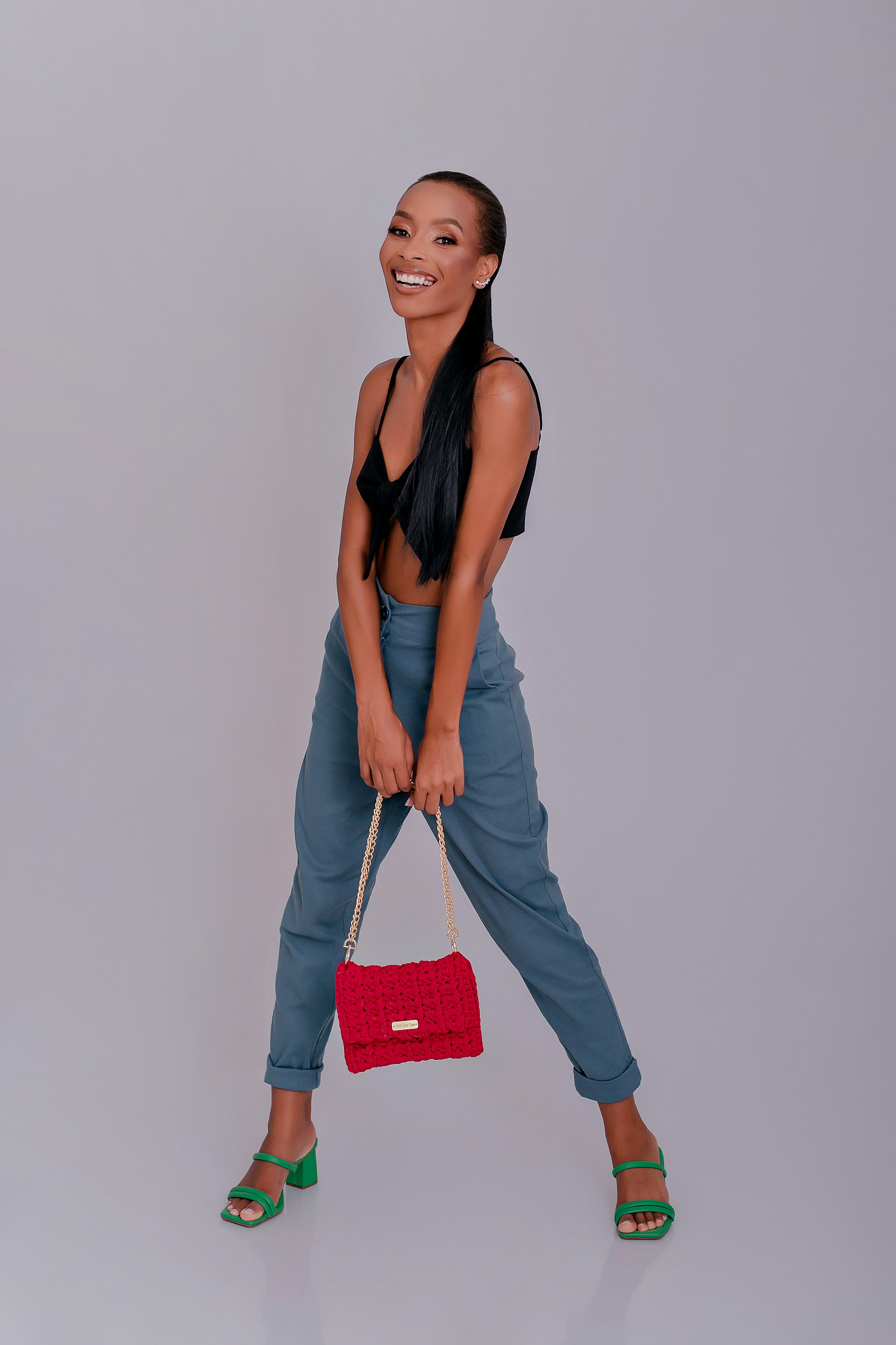 a young black woman with long black hair tied in a ponytail, is smiling confidently while posing in a studio shot against a grey background. she is wearing a black crop top, loosefitting blue pants, and green highheeled sandals. she carries a bright red handbag with a gold chain strap. her pose is relaxed and stylish. the overall image conveys a sense of fashion and confidence.