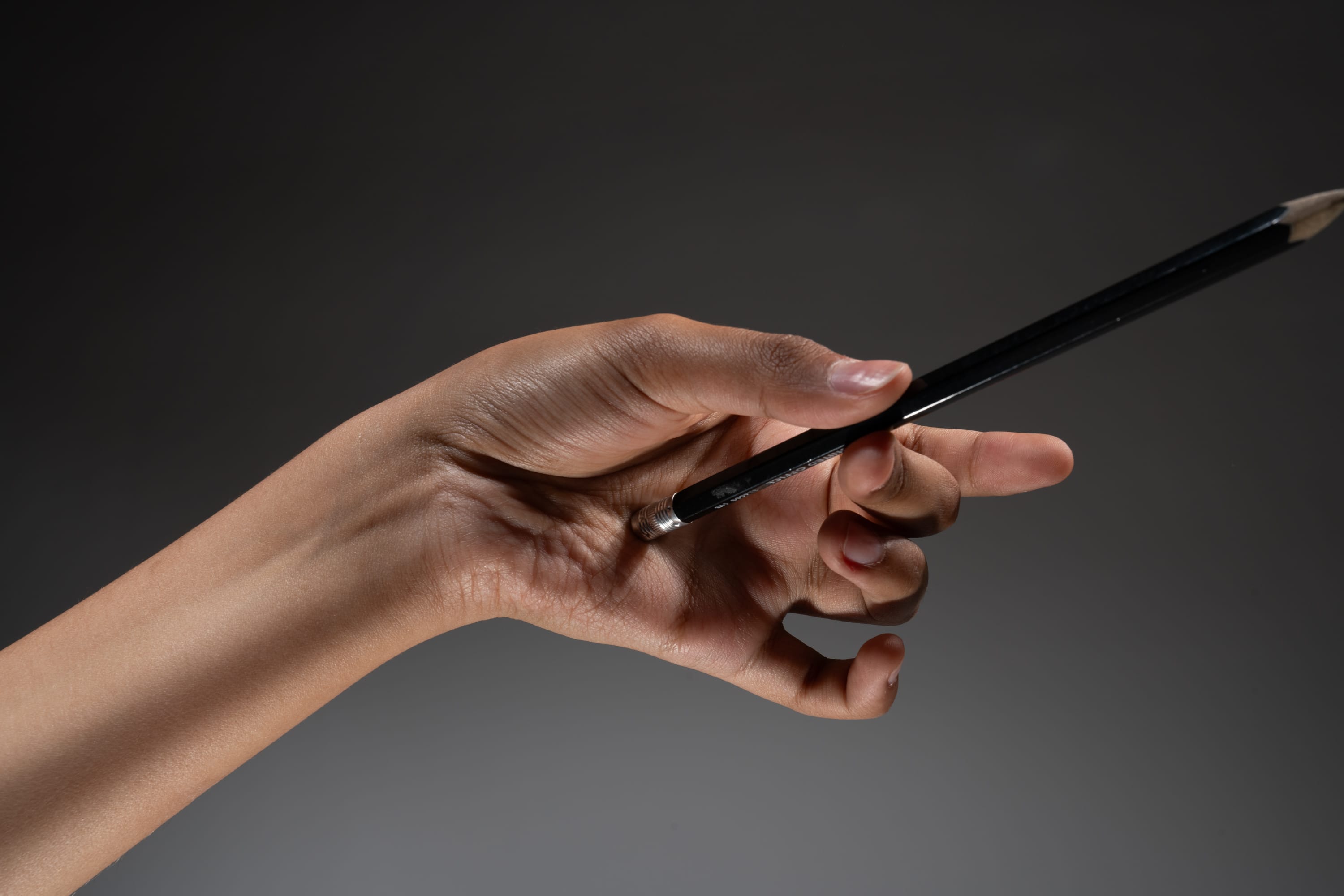 a hand holds a black pencil and points to the right against a dark gray background.