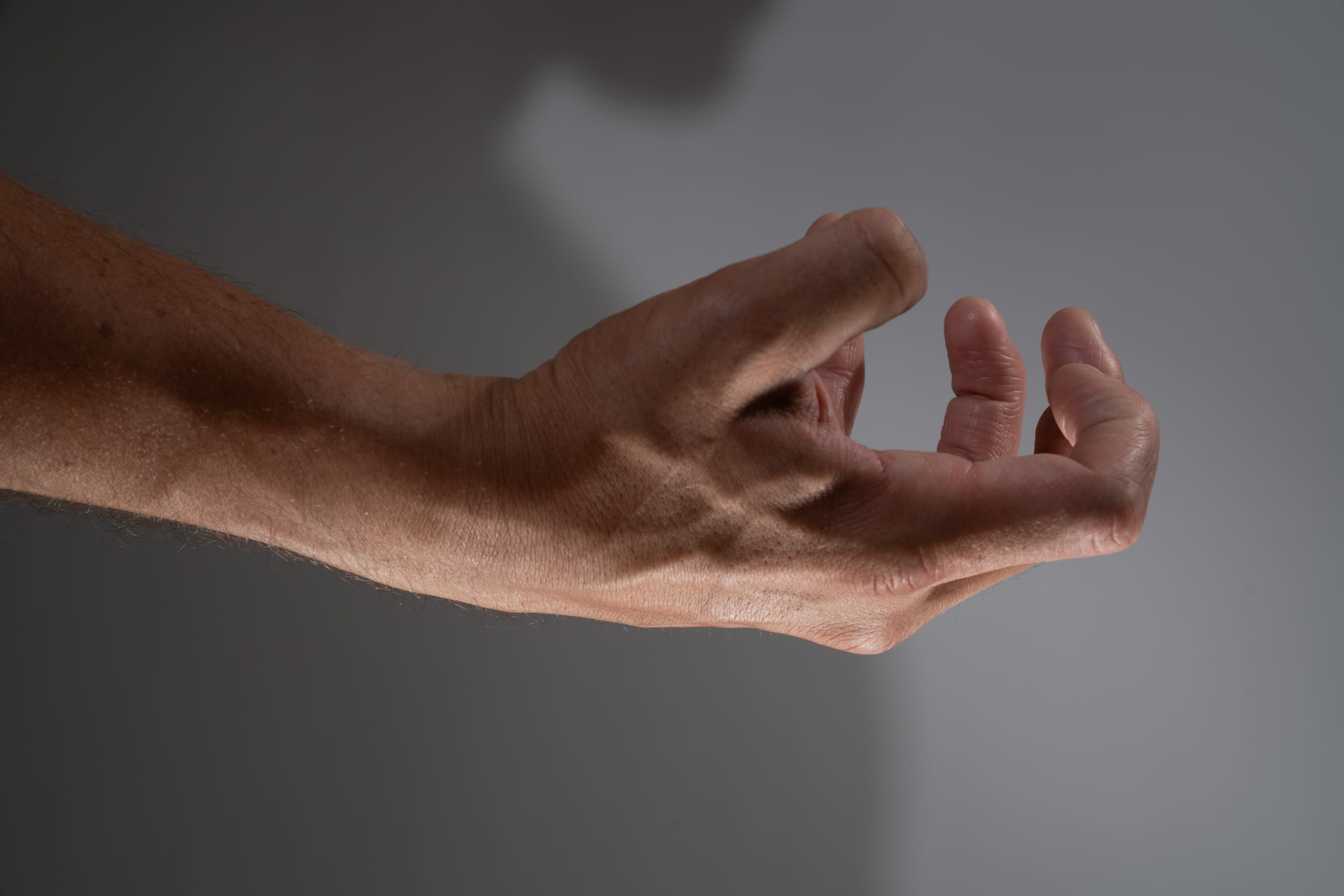 a closeup view of a man's arm and hand extended forward, set against a neutral gray background. the hand is partially clenched, with the fingers curled inward. the image highlights the details of the skin, veins, and shadows created by studio lighting.