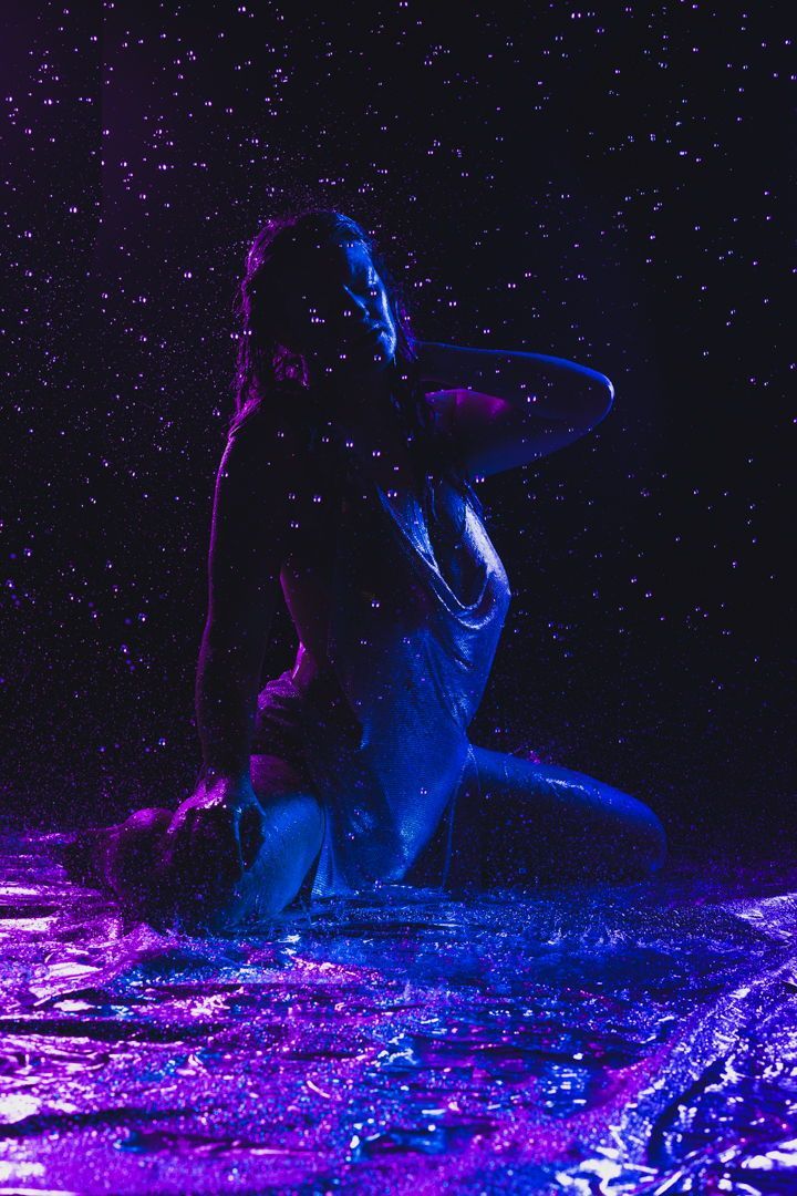 the image depicts a woman, seemingly kneeling, drenched in water under a shower or in the rain. she's wearing a shimmering, wet dress that clings to her body. the scene is set against a dark background with a dramatic use of blue and purple neon lighting that creates a vibrant, almost otherworldly atmosphere.  water droplets are visible in the air around her, adding to the sense of movement and wetness. the overall effect is artistic and sensual, with a focus on the interplay of light, water, and the model's form. the reflective surface she is on enhances the mood and color.