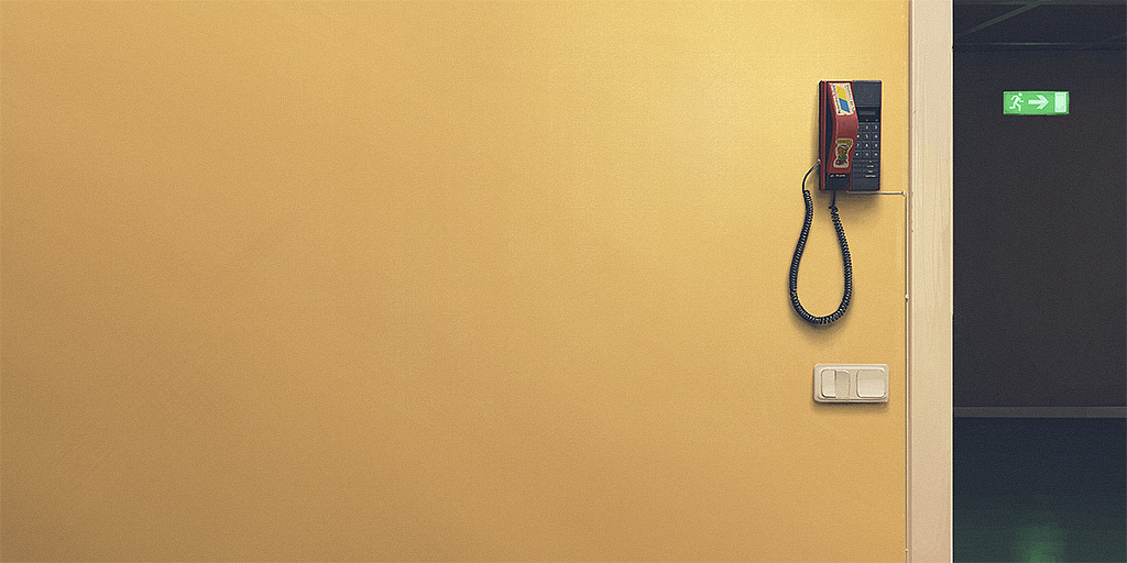 a red phone hangs on a yellow wall next to a white door. the phone has a black cord that hangs down. on the wall is a white light switch. there is a doorway on the right of the image. there is a green exit sign on the door.  the phone is red and black. the wall is a light yellow. the door is white. the exit sign is green. the light switch is white. the cord is black.  the doorway is black.