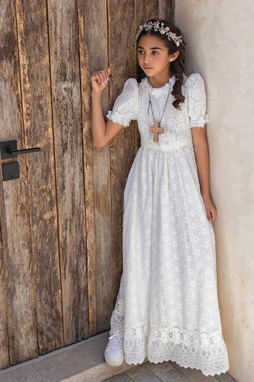 a girl wearing a white communion dress, flower crown, cross necklace, and white sneakers is standing in front of a wooden door and a wall. she is leaning against the wall.